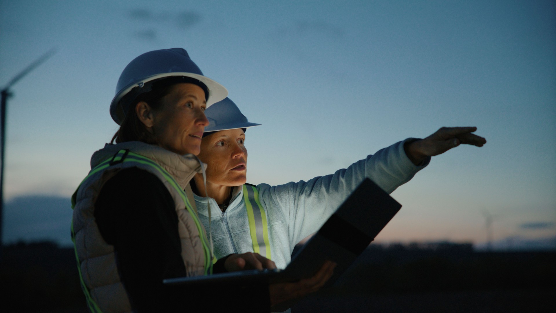 Two Female Engineers in Work Attire with Laptop Discussing About Wind Turbines in Farm at Dusk